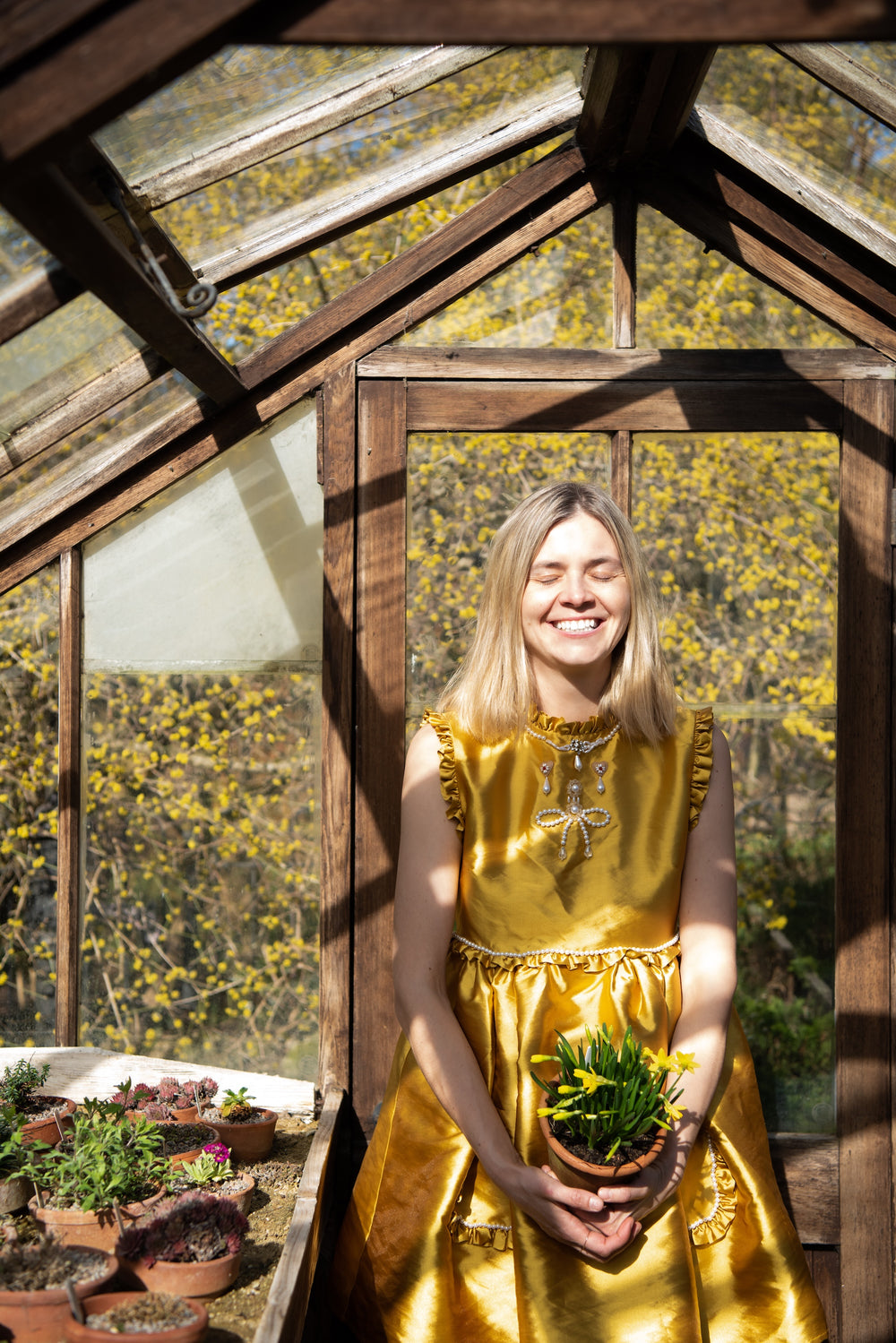 Woman in a gold dress holding a plant inside a greenhouse with yellow flowers outside.