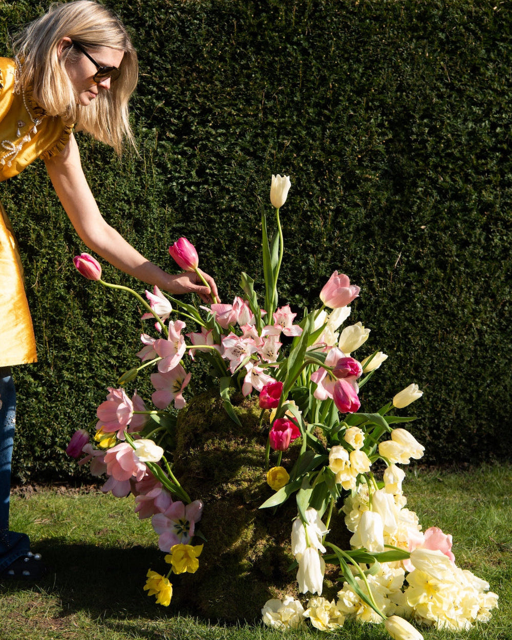 Woman arranging flowers outdoors with a green hedge background